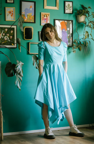 Woman wearing the Bubbles Dress by Cori Tailor — light blue dress with puff sleeves, square neckline, and handkerchief hem, photographed indoors against a teal wall with framed art
