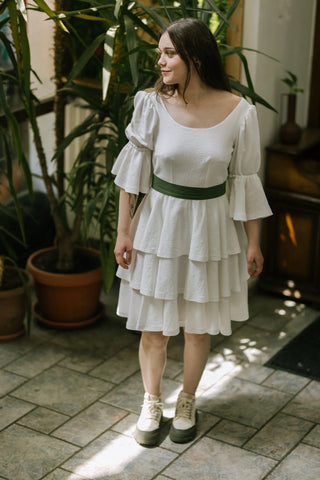 Model in the Freya Dress by Cori Tailor, full front view with puff sleeves, tiered skirt, and green bow belt, posed indoors among plants