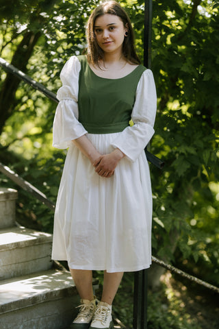 Full front view of Heidi Dress with green bodice and white gathered skirt, model standing on outdoor steps surrounded by greenery.