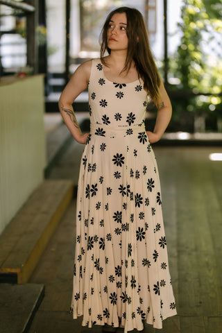 Model wearing the Penny Lane Dress — sleeveless full-length dress with black floral print on soft beige fabric, captured front view while standing on outdoor café terrace.
